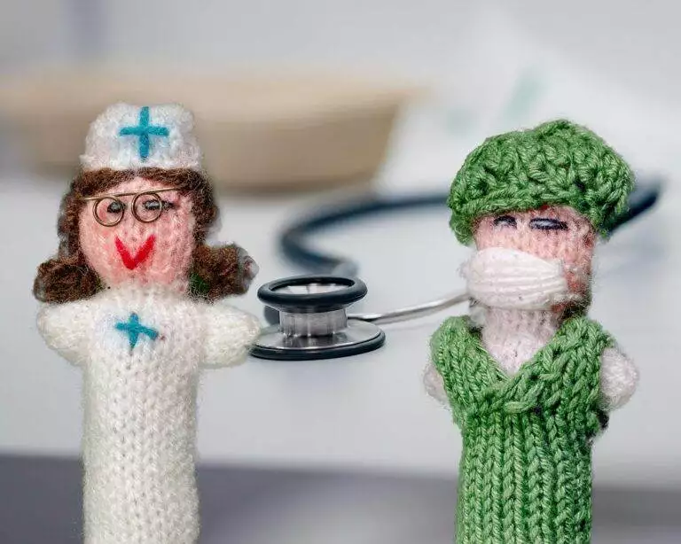 Zwei handgefertigte Strickpüppchen, das eine Ärztin und eine Pflegefachkraft darstellt, mit Stethoskop. Ärztin trägt einen weißen Kittel und die Pflegekraft einen grünen.