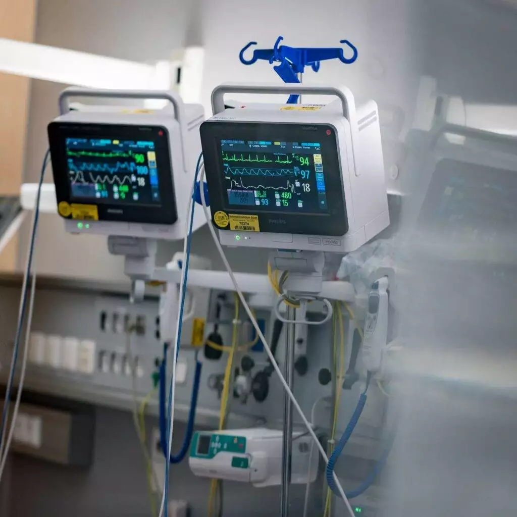Ein Blick in ein Krankenhauszimmer mit zwei Monitoren. Der linke Monitor zeigt die aktuelle Herzfrequenz, den Blutdruck und den Sauerstoffgehalt im Blut eines Patienten.