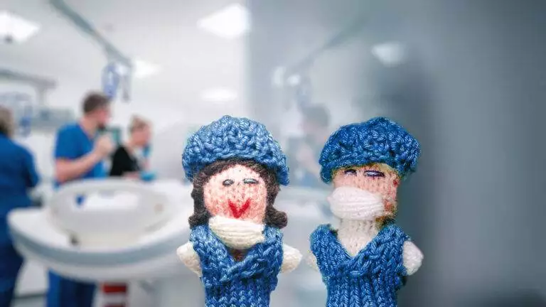 Zwei Strickpüppchen von Pflegekräften in blauen Kitteln befinden sich in einem Zimmer einer Gesundheitseinrichtung, während im Hintergrund echtes Pflegepersonal zu sehen ist.