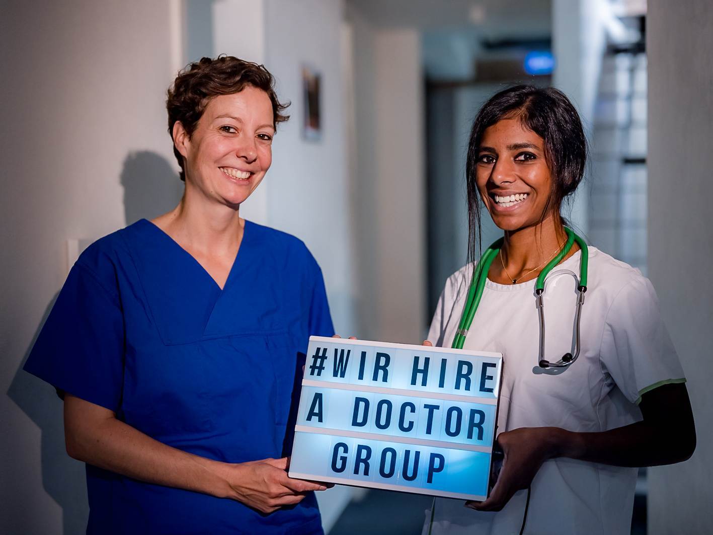 Gesundheits-und-(Kinder)­Krankenpflegerin im blauen Kittel und eine Ärztin im weißen Kittel mit einem Stethoskop um den Hals. Beide lächeln und halten ein Schild mit dem Text: '#WIR HIRE A DOCTOR GROUP.'