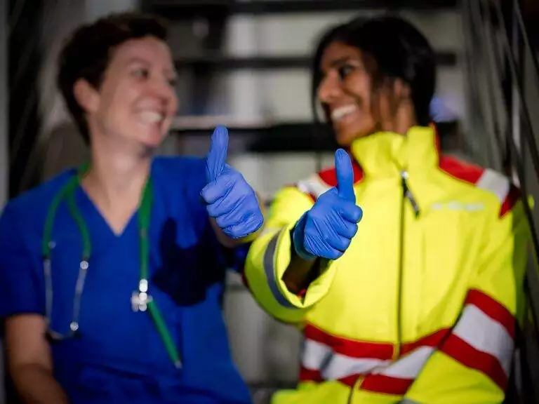 Zwei Frauen, eine in einem weißen Arztkittel mit einem Stethoskop um den Hals und die andere als Rettungskraft. Beide lachen fröhlich, schauen einander an und geben sich gegenseitig Daumen hoch.