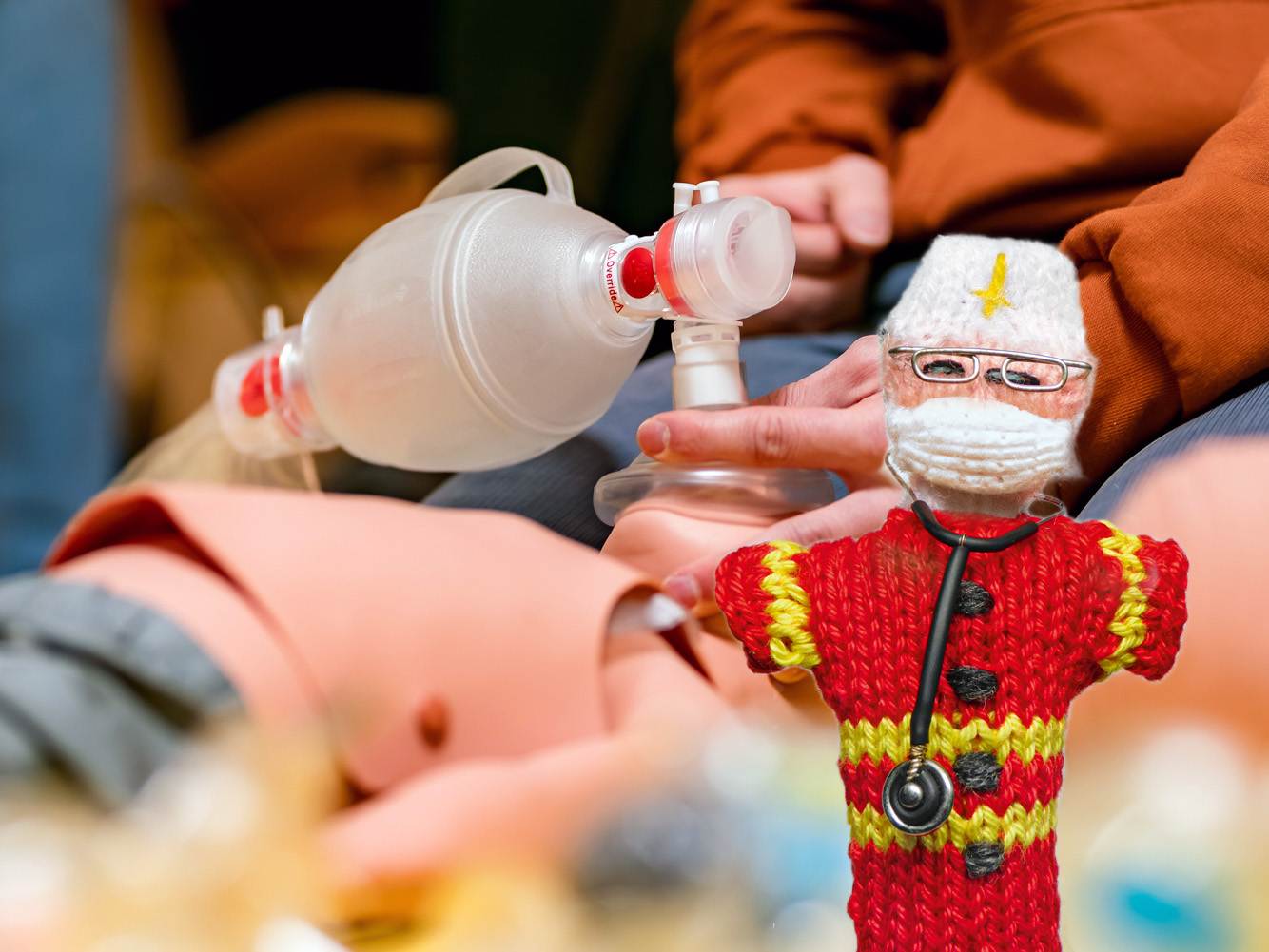 Strickpüppchen eines Kinderarztes in roter Uniform. Im Hintergrund ist ein Training mit Sauerstoffmaske und Kinderpuppen zu sehen.