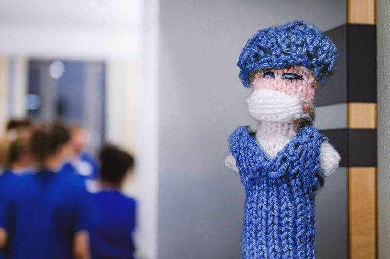 Strickpüppchen in Blau mit Maske: Handgefertigte Pflegekraftfigur mit blauem Strickkleid und Gesichtsmaske, symbolisiert Pflege und Schutz und Prävention Gesundheitseinrichtungen.