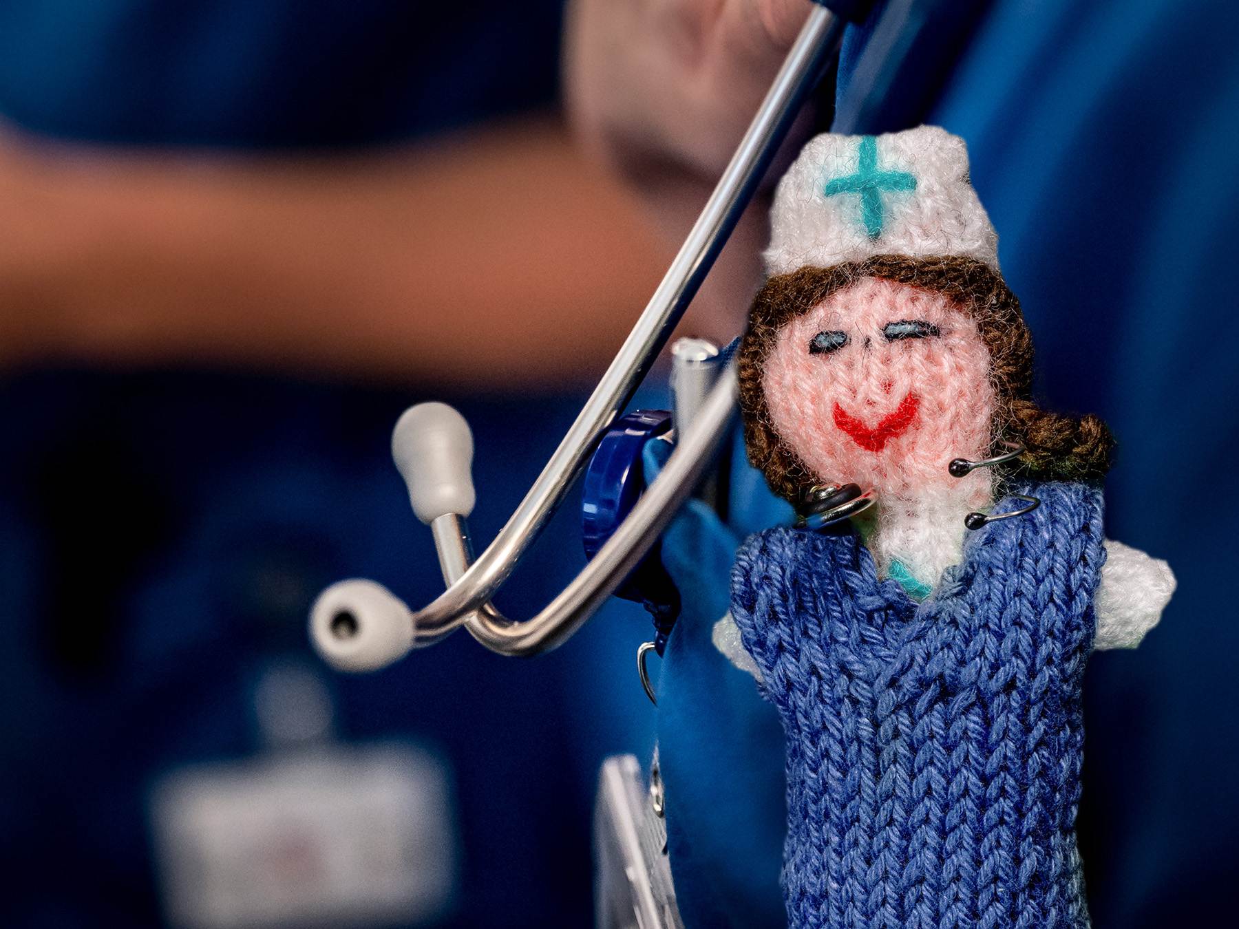 Strickpüppchen einer Gesundheits- und (Kinder-)Krankenpflegerin mit blauem Kittel und einem echten Stethoskop. Der Hintergrund ist bewusst unscharf gehalten und zeigt echte Gesundheits- und (Kinder-)Krankenpfleger.