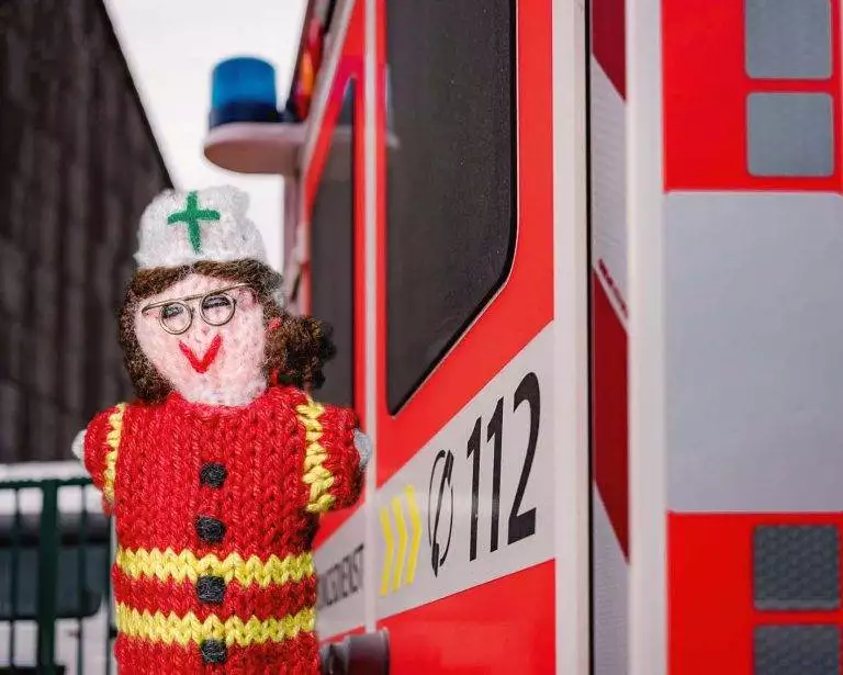 Strickpüppchen einer Rettungskraft in einem roten Kittel neben einem Krankenwagen. Auf dem Krankenwagen steht die Notfallnummer 112.