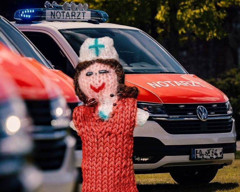 Strickpüppchen einer Notärztin mit roter Uniform. Im Hintergrund sind zwei Rettungswagen zu sehen