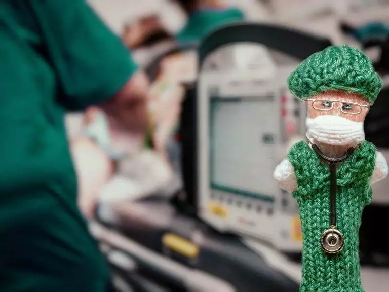 Strickpüppchen einer Anästhesistin im grünen Kittel, im Hintergrund ist ein EKG-Gerät zu sehen.