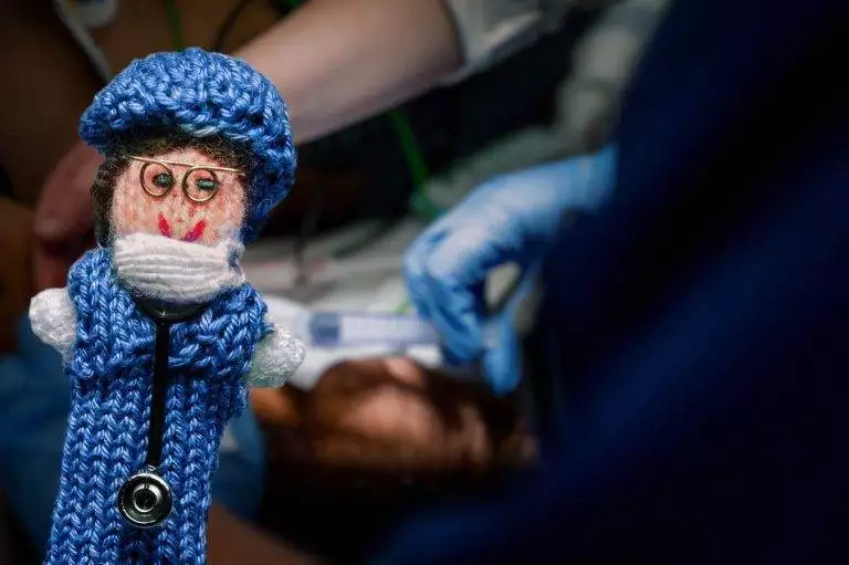 Strickpüppchen einer Pflegekraft in einem blauen Kittel und mit Brille. Im Hintergrund ist ein Schockraum zu sehen