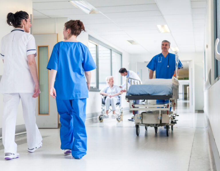 Pflegekräfte in einem Krankenhausflur. Im Vordergrund gehen zwei Pflegekräfte in weißer und blauer Dienstkleidung, während im Hintergrund ein Pfleger mit einem Bett und ein Arzt bei einer Patientin im Rollstuhl zu sehen sind. Der dazugehörige Text handelt von klassischen Arbeitszeitmodellen und Dienstplänen in der Pflege.