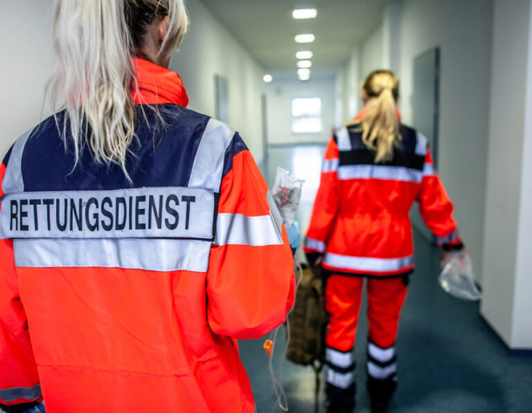 Zwei Rettungskräfte in leuchtend oranger Einsatzkleidung mit der Aufschrift „Rettungsdienst“ auf dem Rücken gehen durch einen Krankenhausflur und tragen medizinische Ausrüstung. Bild von Hire a Paramedic und medilearn