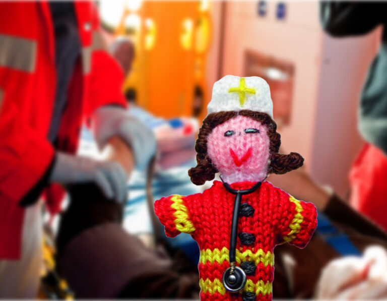 Im Vordergrund steht ein gestricktes Puppenmodell in einem roten Rettungskittel, das einen RettSan darstellt. Im Hintergrund sind Rettungssanitäter und ein Notarzt bei der Patientenversorgung zu sehen. Das Bild stammt von HAP und medi-learn.
